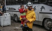 Muhalif savaşçıları ve ailelerini Şam'dan getiren konvoyun önündeki bir Beyaz Miğferler üyesi, kucağında bir çocukla. Mayıs 2018. (© picture-alliance/dpa)